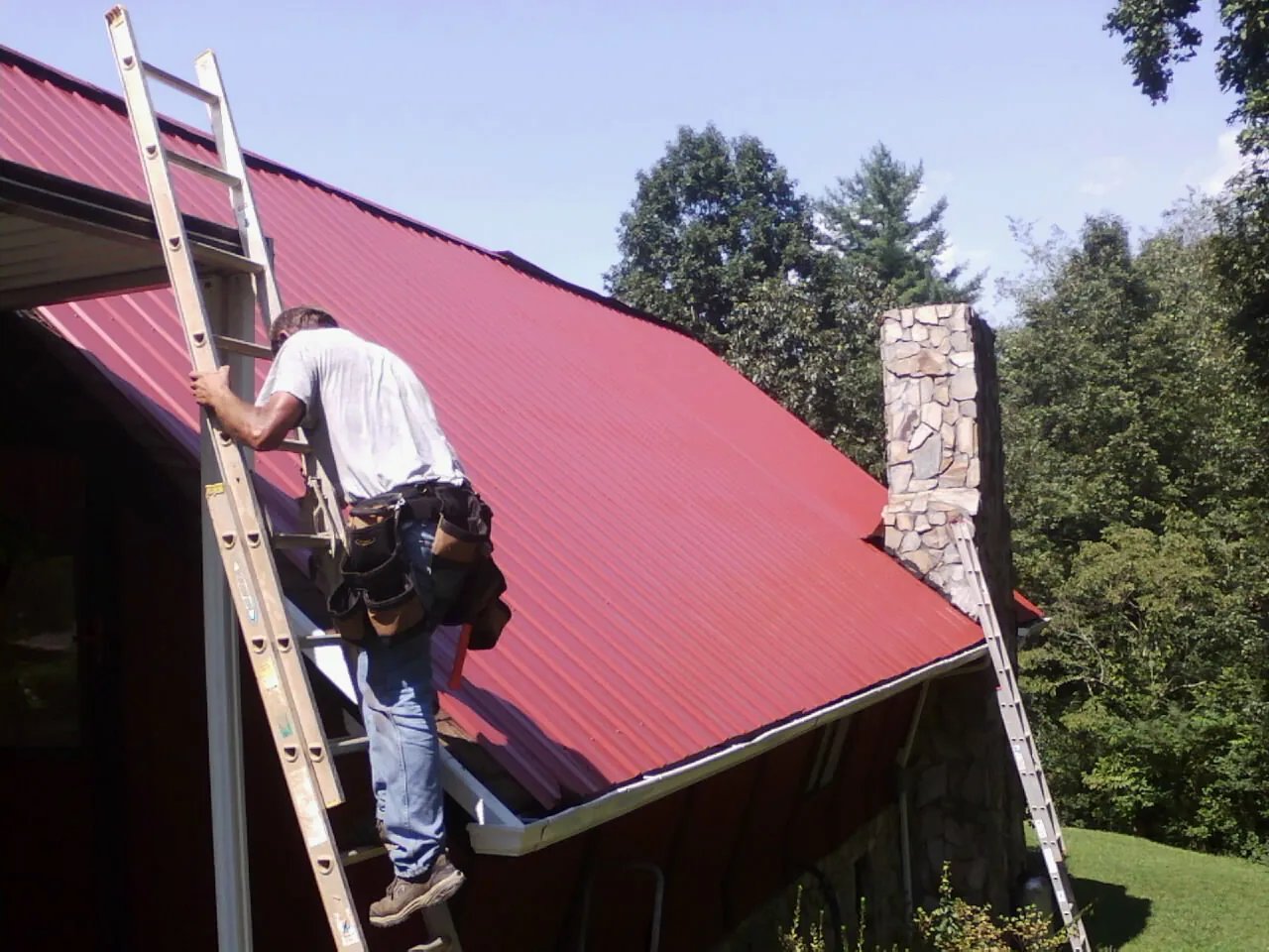 Roofer on a ladder beside a red metal roof and stone chimney — Soffit & Fascia Repair by Roofing Brothers in Brodheadsville